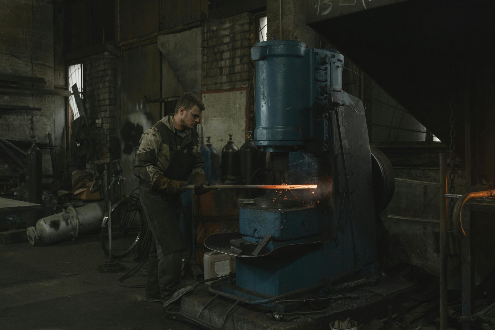 A blacksmith efficiently works with hot metal using industrial machinery in a dimly lit workshop.